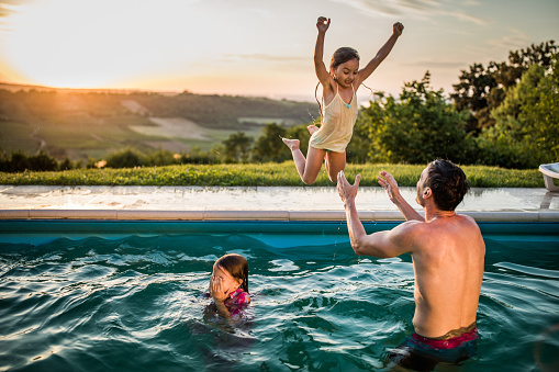 Playful girls having fun with their father in the swimming pool at sunset. One of them is jumping to his father in the pool.
