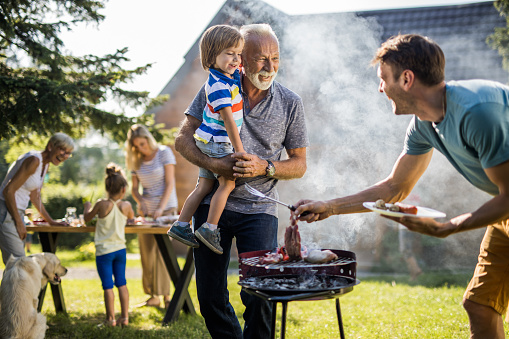 Happy grandfather and grandson talking to young father preparing barbecue in the backyard.