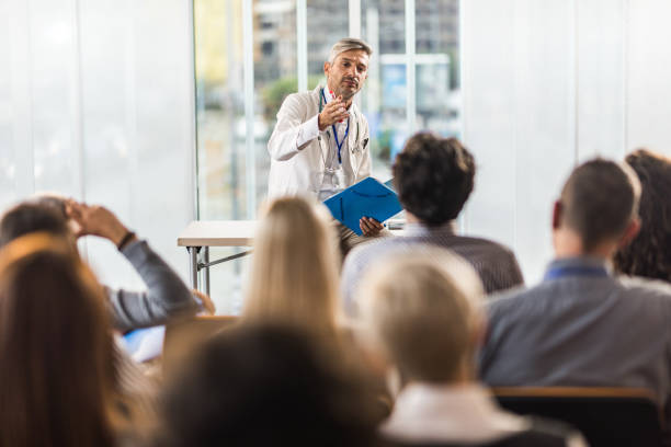 Mid adult doctor teaching on a seminar in a board room. Male doctor talking to large group of people on a seminar in a board room. medical education stock pictures, royalty-free photos & images