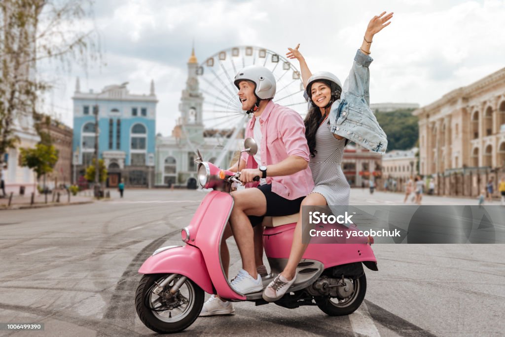 Optimistic jolly couple riding in city Funny ride. Joyful charming man conducting motorbike and woman putting hands up Motor Scooter Stock Photo