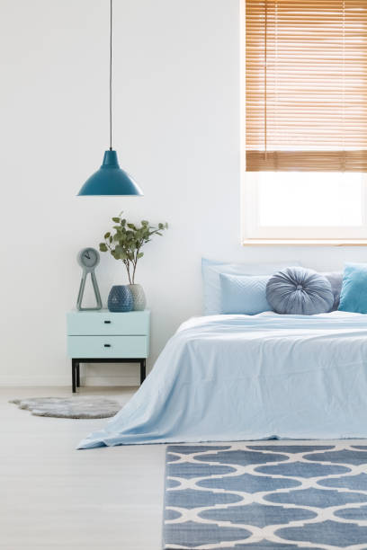 istock White bedroom interior with blue bedding on double bed, navy lamp, bedside table with clock and plant and carpet on the floor 1006515380