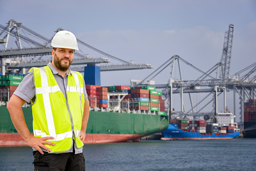 Dockworker standing in front of a container terminal in port. The man is wearing protective workwear and looking into the camera.