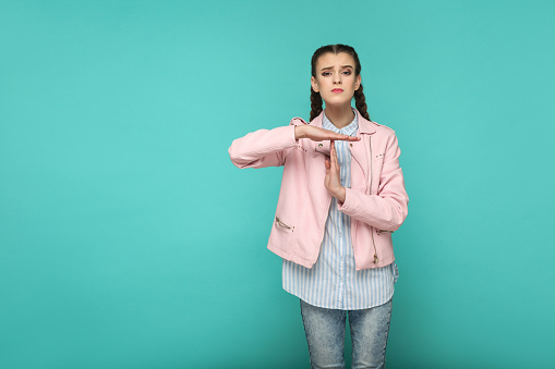 please give me more time. portrait of beautiful cute girl standing with makeup and brown pigtail hairstyle in striped blue shirt pink jacket. indoor, studio shot isolated on blue or green background.