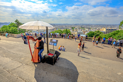 Paris, France - July 3, 2017: skyline above Paris and harp player on the steps of Sacre Coeur de Montmartre. Sacred Heart is a popular tourist landmark. Street artist playing an acoustic instrument.