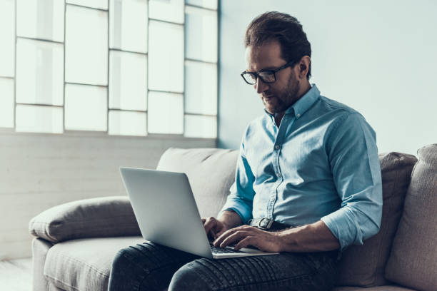 istock Bearded Man in Glasses with Laptop on Gray Sofa. 1006542864