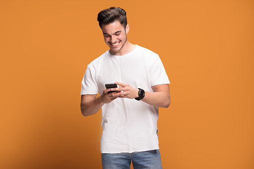 Photo of happy handsome man holding mobile phone in hands, texting. Young guy with big toothy smile posing on yellow background wearing white t-shirt.