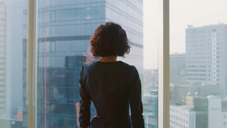 Following Shot of the Successful Businesswoman in a Stylish Dress Walking Through Her Office and Looking out of the Window Thoughtfully. Modern Business Office with Personal Computer and Big City View.