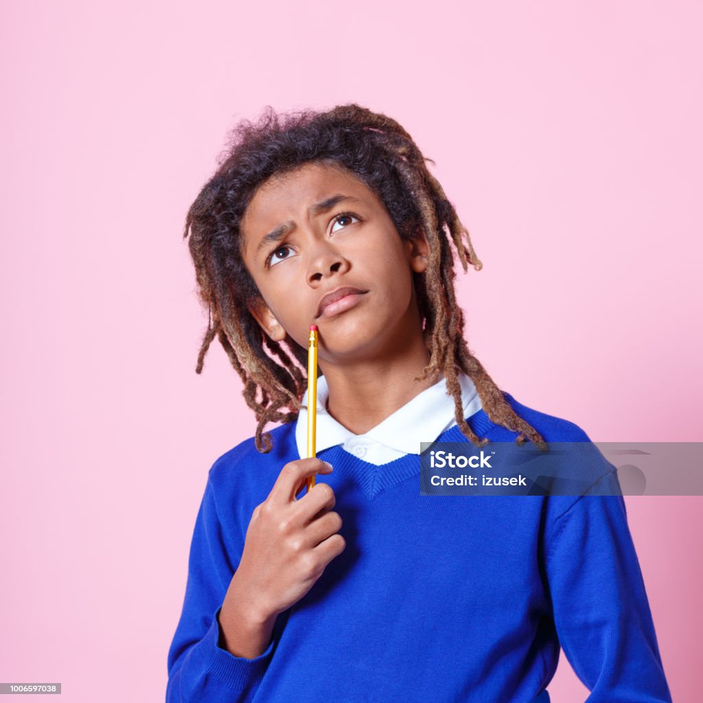 Student looking up and thinking Portrait of young boy in school uniform with pencil looking up and thinking. African school boy with dreadlocks against pink background. Pencil Stock Photo