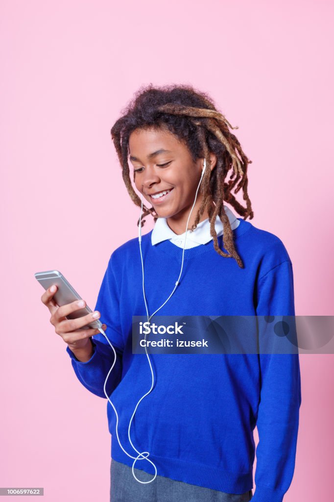 Teen student listening music with phone Teen student playing on mobile phone and listening to music with earphones. School boy with dreadlocks using mobile phone and listening music over pink background. Colored Background Stock Photo