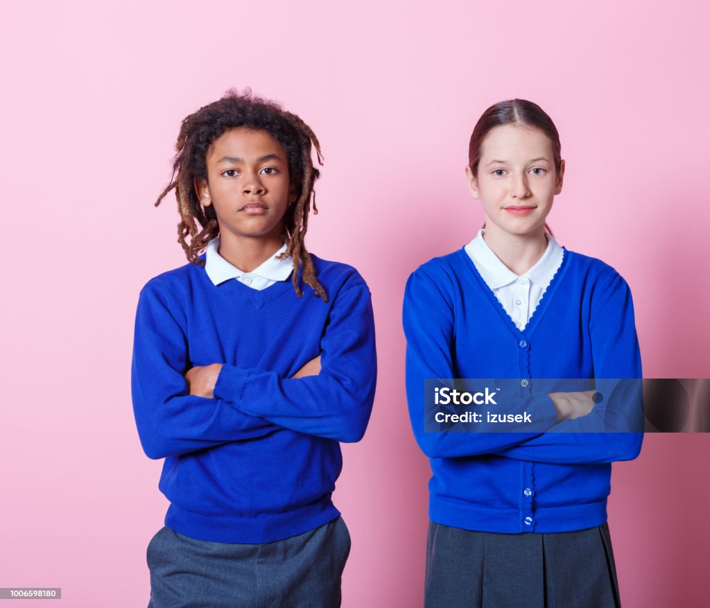 Students standing with arms crossed Two teenage students standing together with their arms crossed. Teenage boy and girl in school uniform looking at camera with blank expression on pink background. Girls Stock Photo