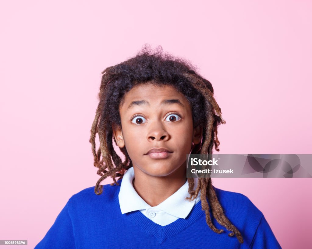 School boy looking surprised Close up portrait of young boy with dreadlocks looking at camera with his eyes wide open. Surprised afro american school boy against pink background. Surprise Stock Photo