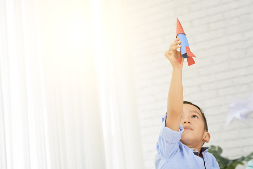 Young Asian boy holding rocket on raised hand standingÂ in art class in backlit