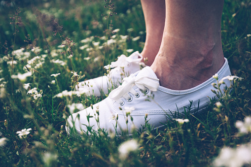 Woman in white sneakers standing on spring meadow with flowers. Feet close-up. Vintage.