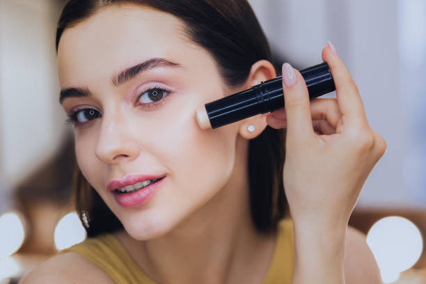 charming woman using concealer stick while putting makeup on - make up imagens e fotografias de stock
