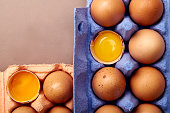 Horizontal closeup shot with two colorful cardboard containers of pink and violet color with chicken eggs, two egg yolks open and visible.