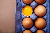 Horizontal closeup shot with dozen of chicken eggs in colorful violet cardborad container, one egg yolk visible