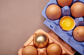 Horizontal closeup shot with two colorful cardboard containers of pink and violet color with chicken eggs,  one yolk visible, one egg painted golden. Easter concept