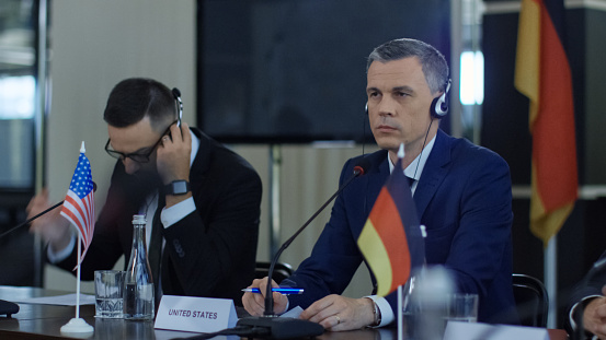 Representative men of different countries sitting at table on conference and listening to speech interpretation in headphones