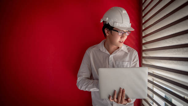 istock Young Asian male engineer or architect wearing protective helmet and glasses working with laptop computer with red wall in the background. Architecture, Engineering and building construction concepts 1006696252