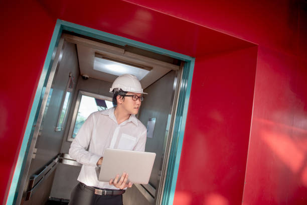 istock Young Asian male engineer or technician worker holding laptop computer in opened elevator door that surrounded by red wall. Mechanical engineering or building construction and maintenance concepts 1006696262