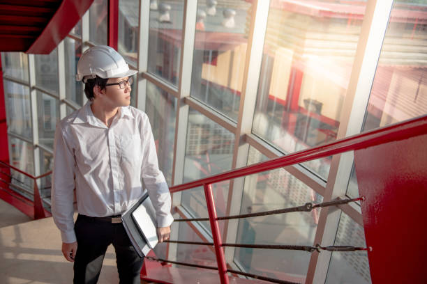 istock Young Asian male engineer or architect holding files wearing protective safety helmet on red stair at construction site. Civil engineering, Architecture and building construction concept 1006696298