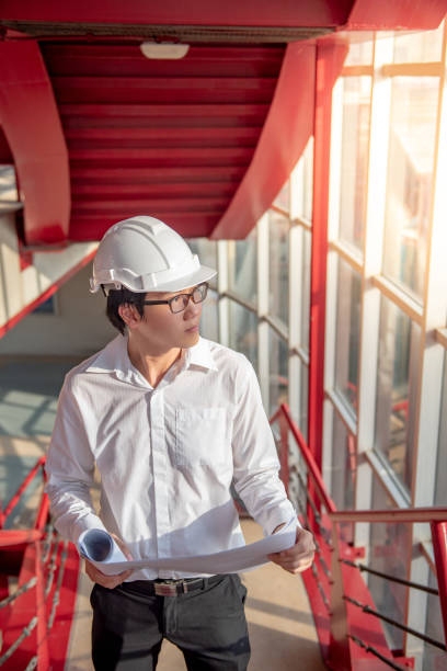 istock Young Asian male engineer or architect holding blueprints wearing protective safety helmet on red stair at construction site. Civil engineering, Architecture and building construction concept 1006696372