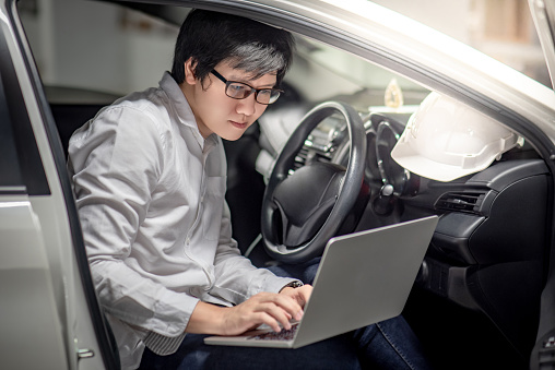 Young Asian male mechanical engineer working with laptop computer while sitting with white safety helmet in the car. Mechanic engineering concepts