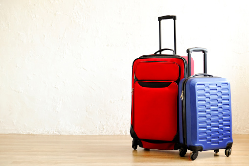 Red textile suitcase & blue hard shell luggage with extended telescopic handle up on wooden floor, white wall background. Couples retreat trip concept. Packed traveling baggages. Close up, copy space.