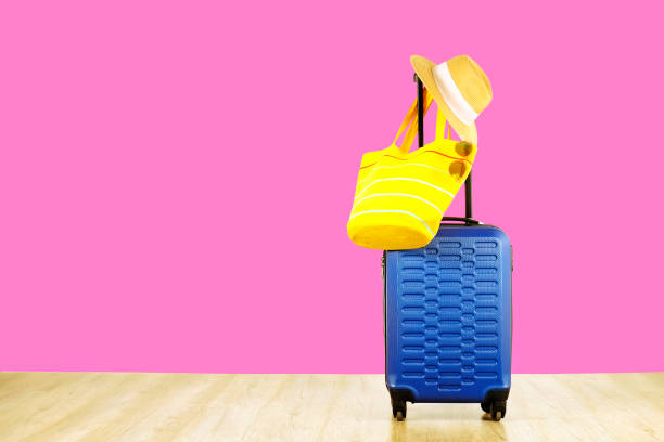 istock Single blue plastic hard shell luggage with women's straw hat hanging on extended telescopic handle, yellow beach bag, sunglasses. One suitcase prepared for vacation trip. Pink background, copy space 1006741410