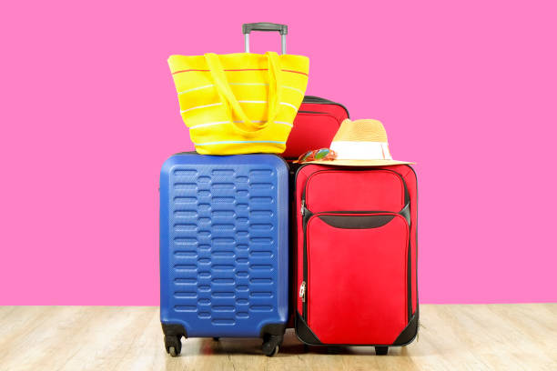 istock Three suitcases, red textile & blue hard shell luggage, extended telescopic handle, straw hat, beach bag, mirrored sunglasses, pink wall background. Family trip concept. Close up, copy space 1006742038