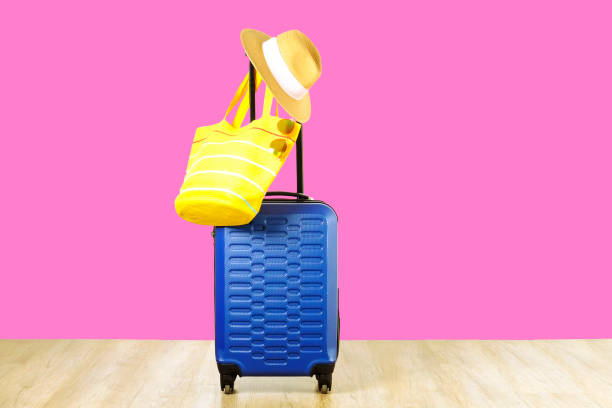 istock Single blue plastic hard shell luggage with women's straw hat hanging on extended telescopic handle, yellow beach bag, sunglasses. One suitcase prepared for vacation trip. Pink background, copy space 1006742048
