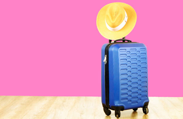 istock Single blue plastic hard shell luggage with woman straw hat hanging on extended telescopic handle. One suitcase. Traveling alone concept. Bright pink wall background, copy space for text, close up. 1006744170