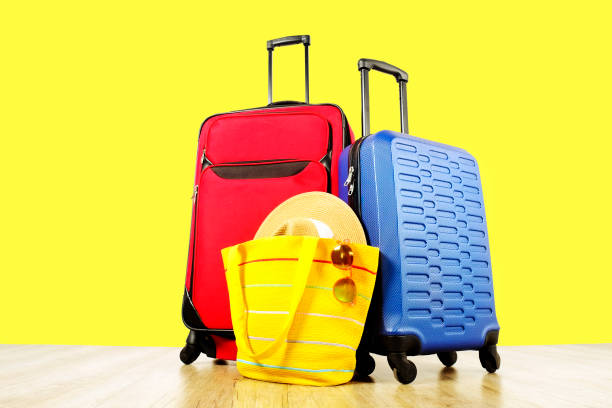 Red textile suitcase & blue hard shell luggage, extended telescopic handle, straw hat, yellow beach bag, mirrored sunglasses, yellow wall background. Couples retreat trip concept. Close up, copy space