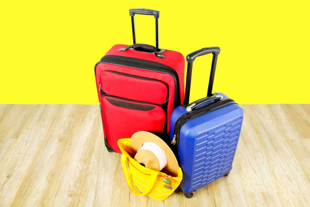 istock Red textile suitcase & blue hard shell luggage, extended telescopic handle, straw hat, yellow beach bag, mirrored sunglasses, yellow wall background. Couples retreat trip concept. Close up, copy space 1006744270