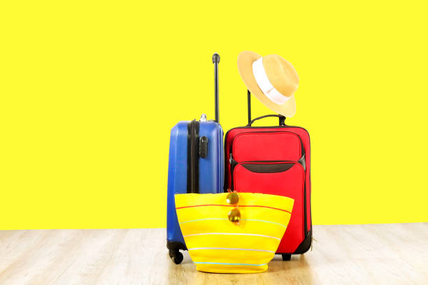 istock Red textile suitcase & blue hard shell luggage, extended telescopic handle, straw hat, yellow beach bag, mirrored sunglasses, yellow wall background. Couples retreat trip concept. Close up, copy space 1006744292