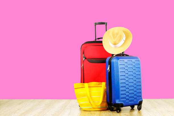 Red textile suitcase & blue hard shell luggage with extended telescopic handle, straw hat, yellow beach bag on wooden floor, pink wall background. Couples retreat trip concept. Close up, copy space