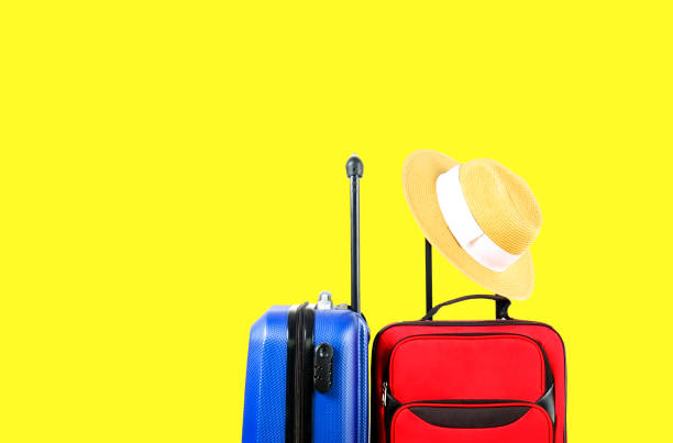 istock Red textile suitcase & blue hard shell luggage with straw hat hanging on extended telescopic handle, bright yellow isolated background. Couples retreat trip concept. Close up, copy space. 1006744340