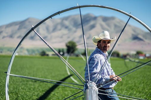Mid aged cowboy  leaning on watering wheel and pipe looking at camera, in the fresh field of crop  Outside Salt Lake City, Utah.