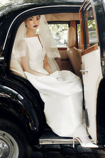 istock gorgeous elegant bride posing in stylish retro black car, sitting inside in saloon.  luxury wedding in vintage style. unusual gown and veil 1006787092
