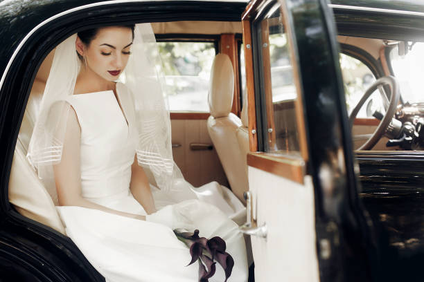 istock gorgeous elegant bride posing in stylish retro black car, sitting inside in saloon holding bouquet of callas. luxury wedding in vintage style. unusual gown and veil 1006787612