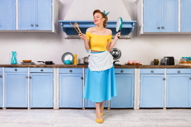 Retro pin housewife sitting in the kitchen Retro pin up girl woman female housewife wearing colorful top, skirt and white apron and yellow high heels holding wooden spoons and pan standing in the blue kitchen with blue cabinets and utensils. pin up girl photos stock pictures, royalty-free photos & images