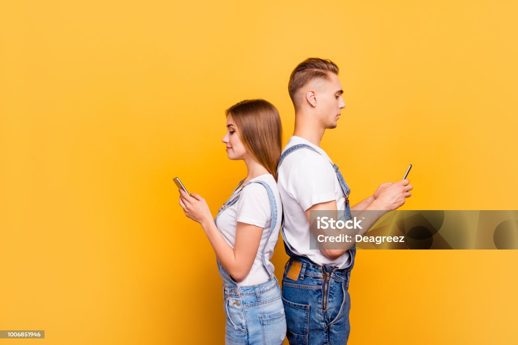 Portrait of cute young students couple wearing casual, standing back to back, holding their smartphones in hands, seriously looking at screens, over yellow background, isolated, copyspace Mobile Phone Stock Photo