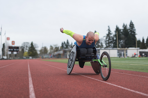 A Caucasian para athlete trains at a stadium track in his wheelchair. He is racing toward the camera and has an intense expression.