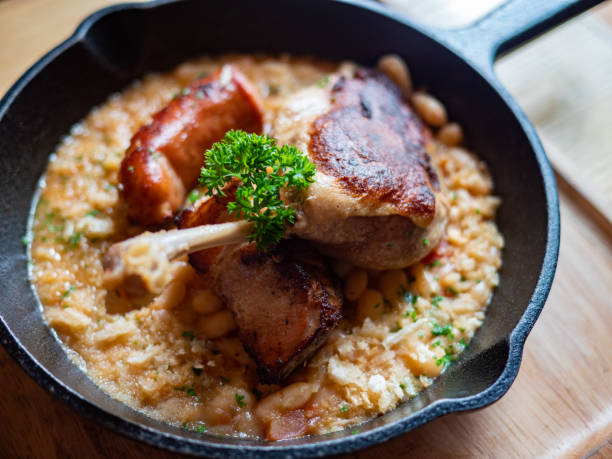 Trio Cassoulet of duck confit, pork belly and smoked sausage on a bed of white beans in a black cast iron pan. Sizzling hot, rich, slow-cooked Trio Cassoulet of duck confit, pork belly and smoked sausage on a bed of white beans in a black cast iron pan on a wooden table. Natural Light. confit stock pictures, royalty-free photos & images