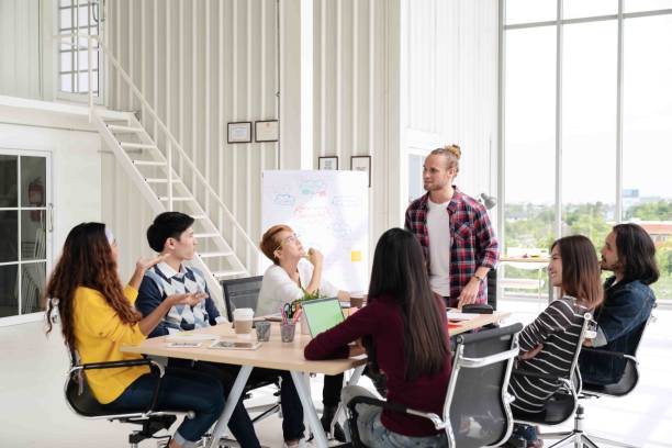 istock Young caucasian creative man standing and making presentation at modern office happy talking and brainstorming with team. Casual multiethnic diverse people business meeting concept with rear view. 1006991594