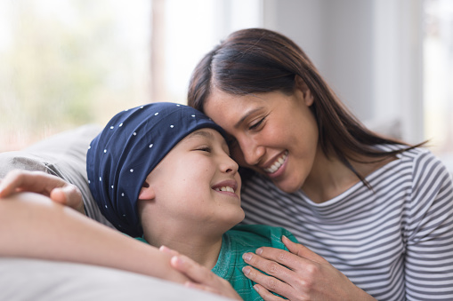 A beautiful ethnic mom embraces her 10-year old son who is battling cancer. Their faces are snuggled up to one another affectionately and they're smiling as they sit on a couch by the window. He is wearing a headcovering.