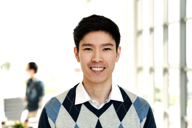 portrait of young attractive asian creative man smiling and looking at camera in modern office feeling confident and positive. headshot of chinese male employee, entrepreneur or college student. - human face chinese ethnicity close up men imagens e fotografias de stock