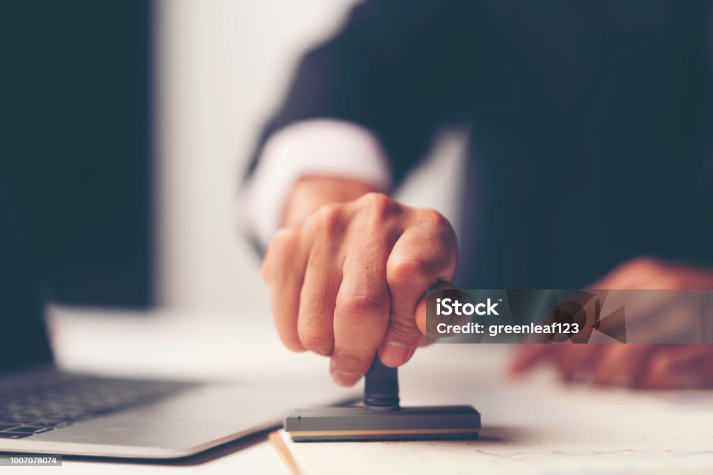 Primo piano della stampa a mano di una persona con timbro approvato sul documento alla scrivania - Foto stock royalty-free di Timbro