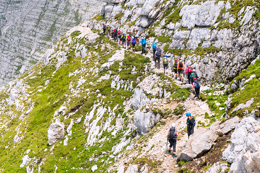 Altopiano del Montasio, Italy- July 14, 2018: A line of hikers on mountain surrounded with slopes on trail to peak Spik Hude police or  Cima di Terrarossavrha,  2420 m high in Italian Alps near border with Slovenia. This is rough terrain the homeland of Alpine ibex and is popular alpine trail but very steep with  the possible fall of stones accidentally triggered by ibex.  This is place to we admire the highest peaks of the western Julian alps.