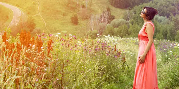 A middle-aged woman in a pink dress in a meadow. The woman looks dreamily at the sky, against the background of the road leading to the top of the hill.